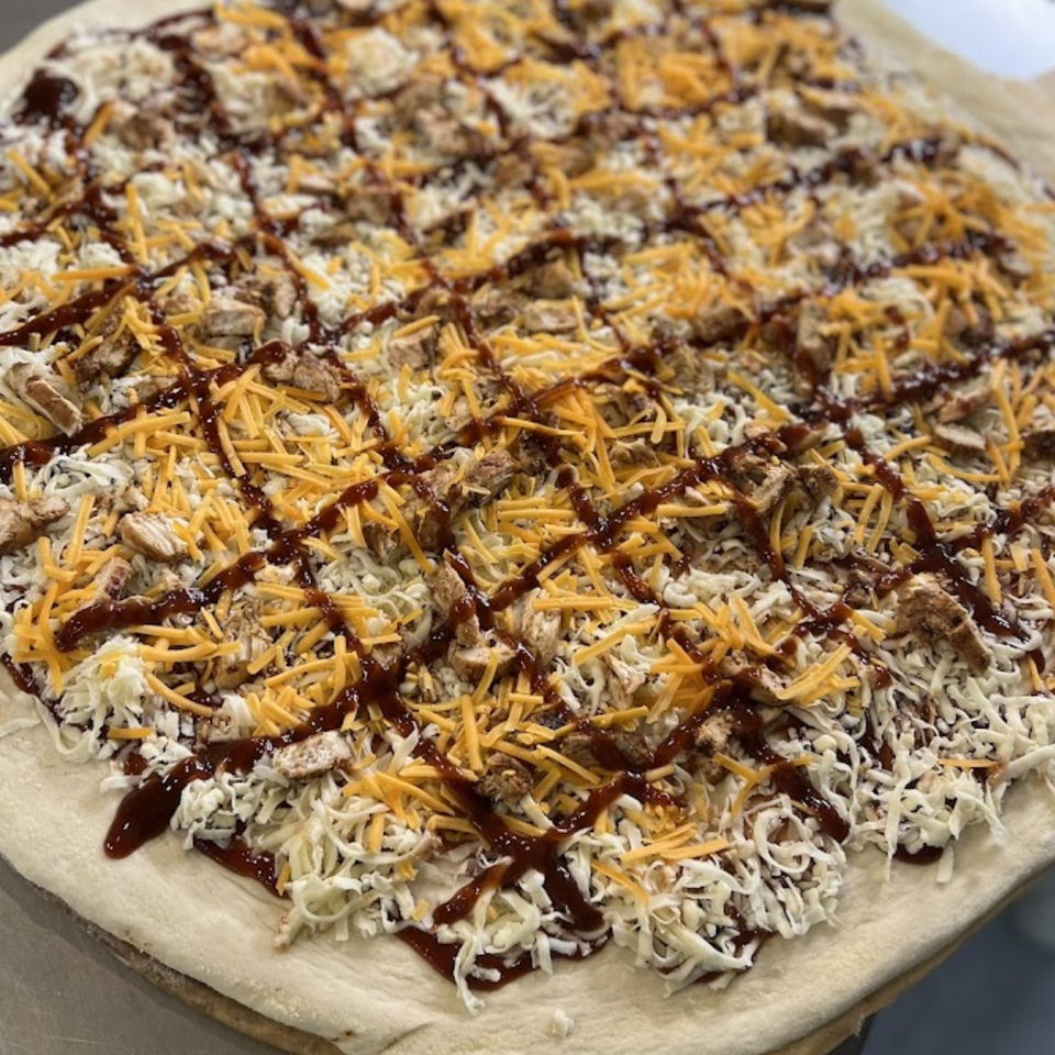 Uncooked Sal's Pizzeria BBQ chicken pizza with shredded mozzarella, cheddar, and BBQ sauce on dough.